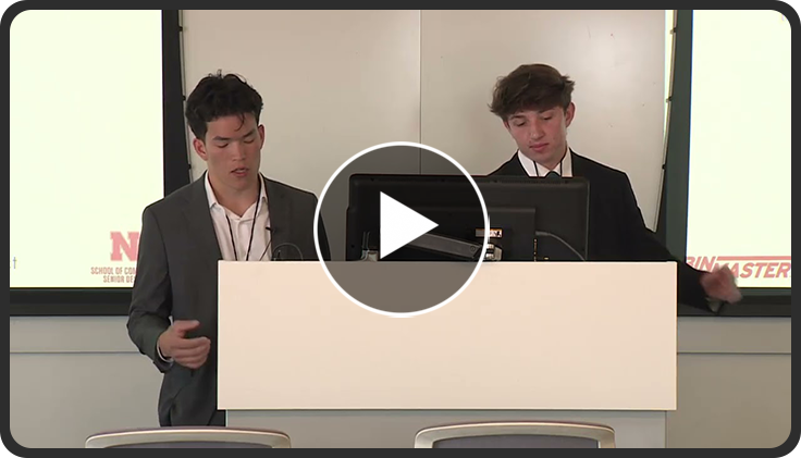 Two speakers stand at a podium in a conference room, presenting slides on two screens.