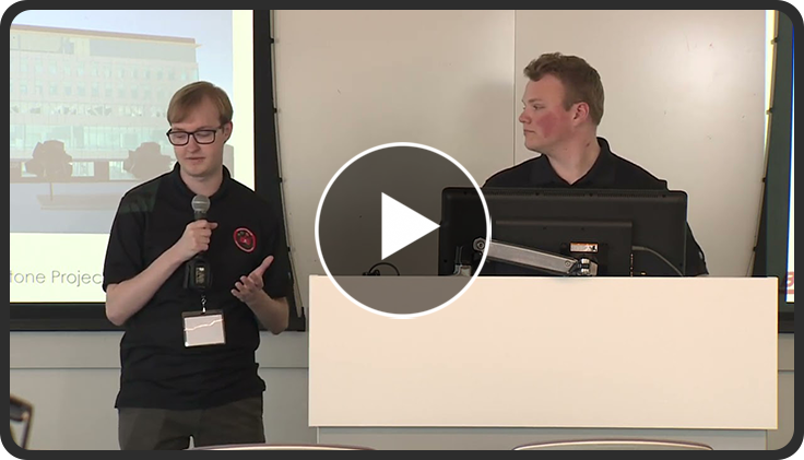 Two individuals presenting in a conference room, one speaking into a microphone.