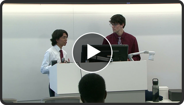 Two people standing at a podium, speaking to an audience in a lecture setting.