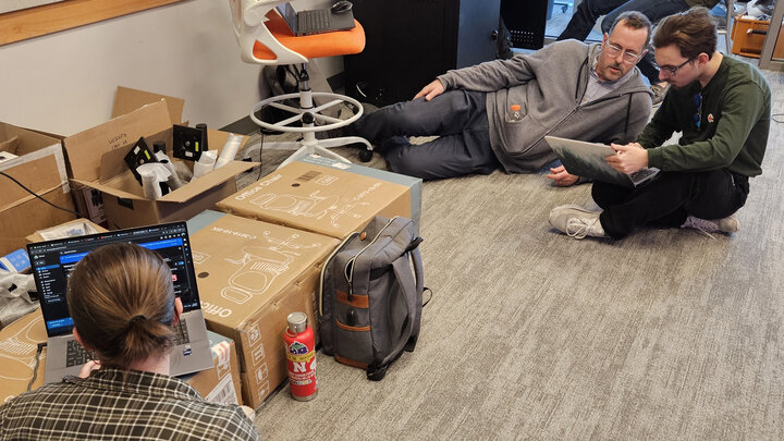 A professor in a grey sweater and jeans talks with a student demonstrating on a laptop wearing glasses and a green shirt.
