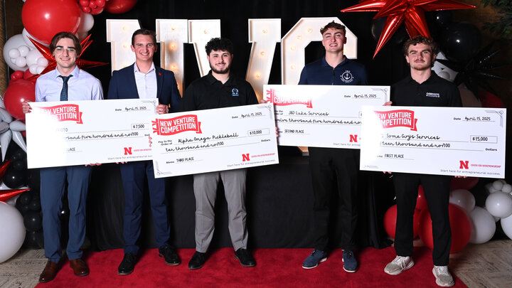 Five young men hold oversized checks in front of the letters "NVC" and red and white balloons.