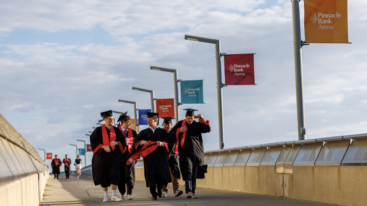 Graduates in black robes and mortar boards with red stoles walk on a pedestrian bridge in the early morning.