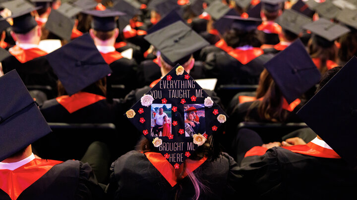 A graduate's decorated mortarboard reads "Everything you taught me brought me here" with two family photos and flowers. The graduate sits among several rows of graduates.