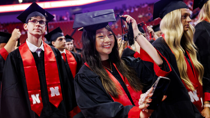 Graduate Christina Dinh records herself on her smartphone as she moves her tassel from right to left. She is wearing a black graduation robe with black mortar board and red stole, near other Husker graduates.