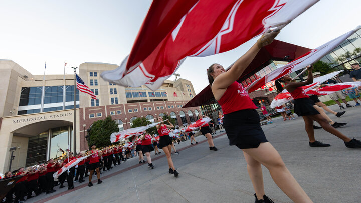 Color guard members — wearing red sleeveless shirts, black skirts and black shoes — wave Nebraska flags as the Cornhusker Marching Band performs in the background on the East Stadium Plaza at Memorial Stadium. The instrumentalists are wearing red polo shirts, black pants and black shoes.
