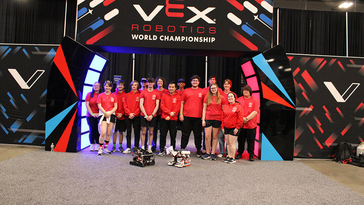 Team in red shirts standing under a VEX Robotics World Championship sign with robots in front.